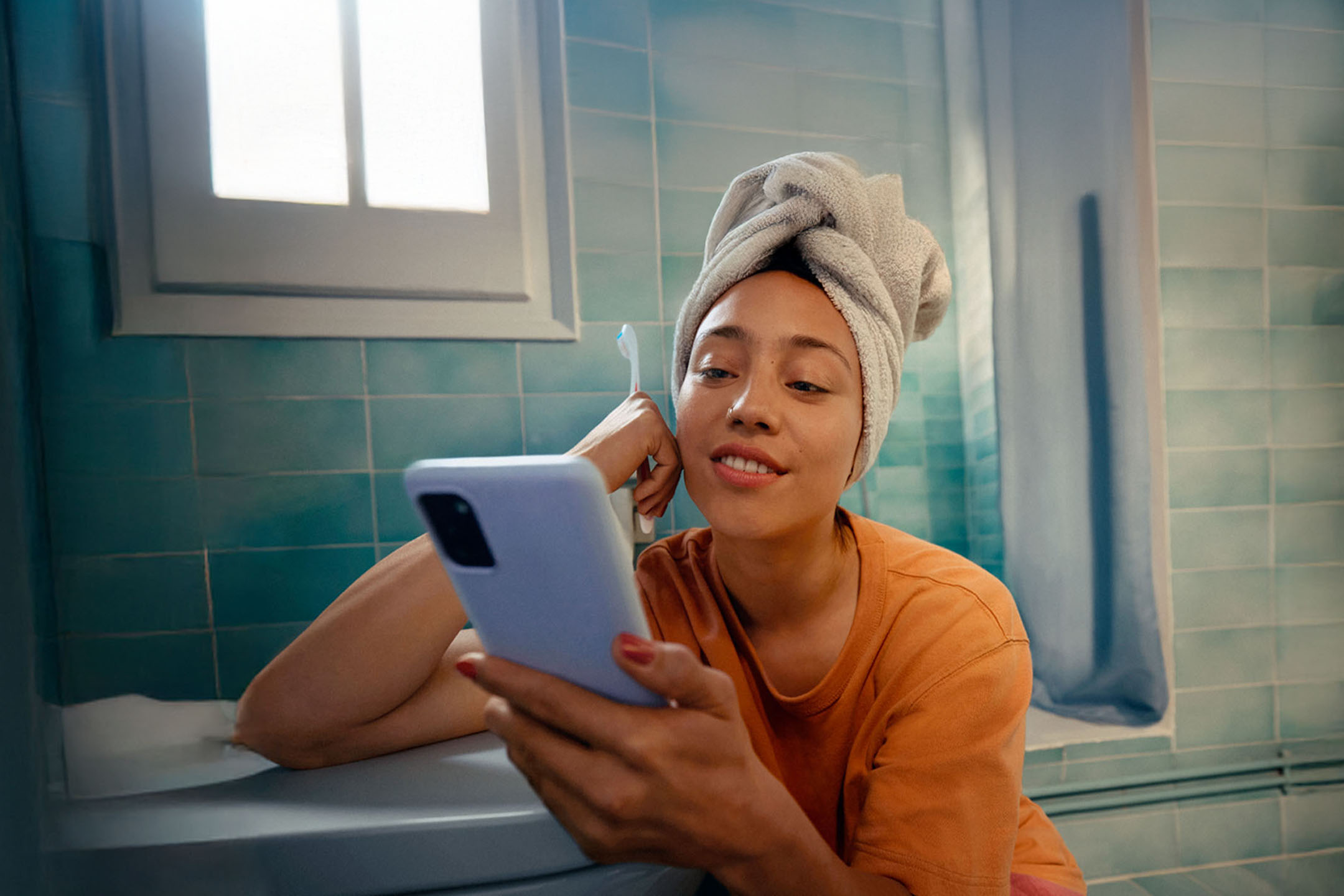 Frau mit Handtuch auf dem Kopf, benutzt ein Smartphone im Badezimmer.