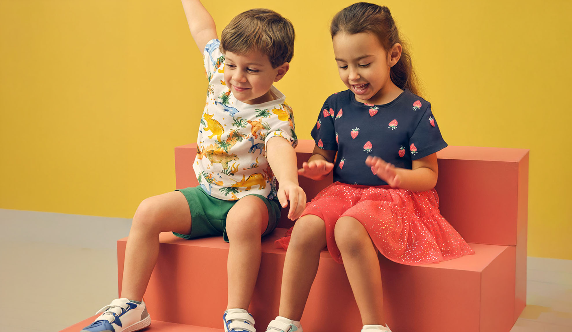 Zwei Kinder in Sommerkleidung, ein Junge im Dinosaurier-T-Shirt und ein Mädchen im Erdbeer-T-Shirt.