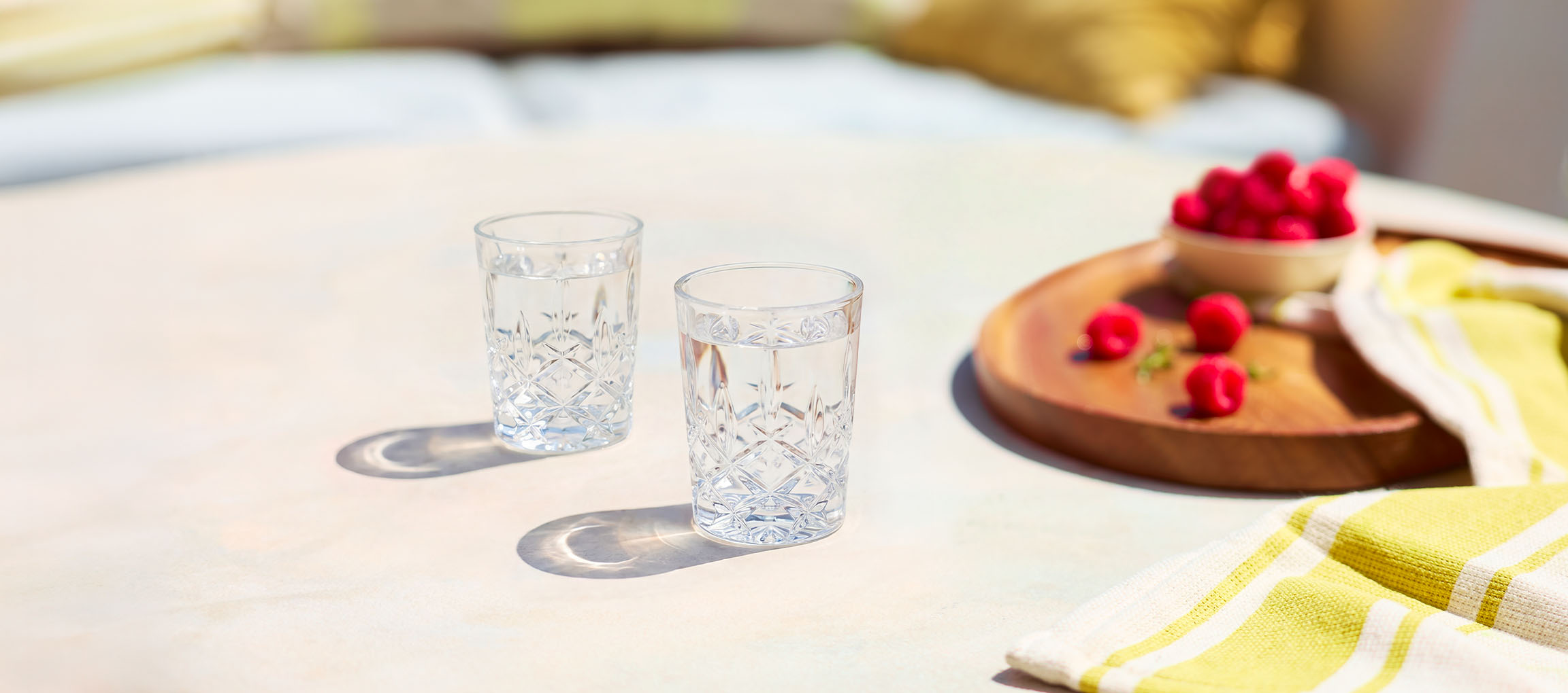 Zwei Gläser Wasser und ein Holztablett mit Himbeeren auf einem Tisch im Freien.