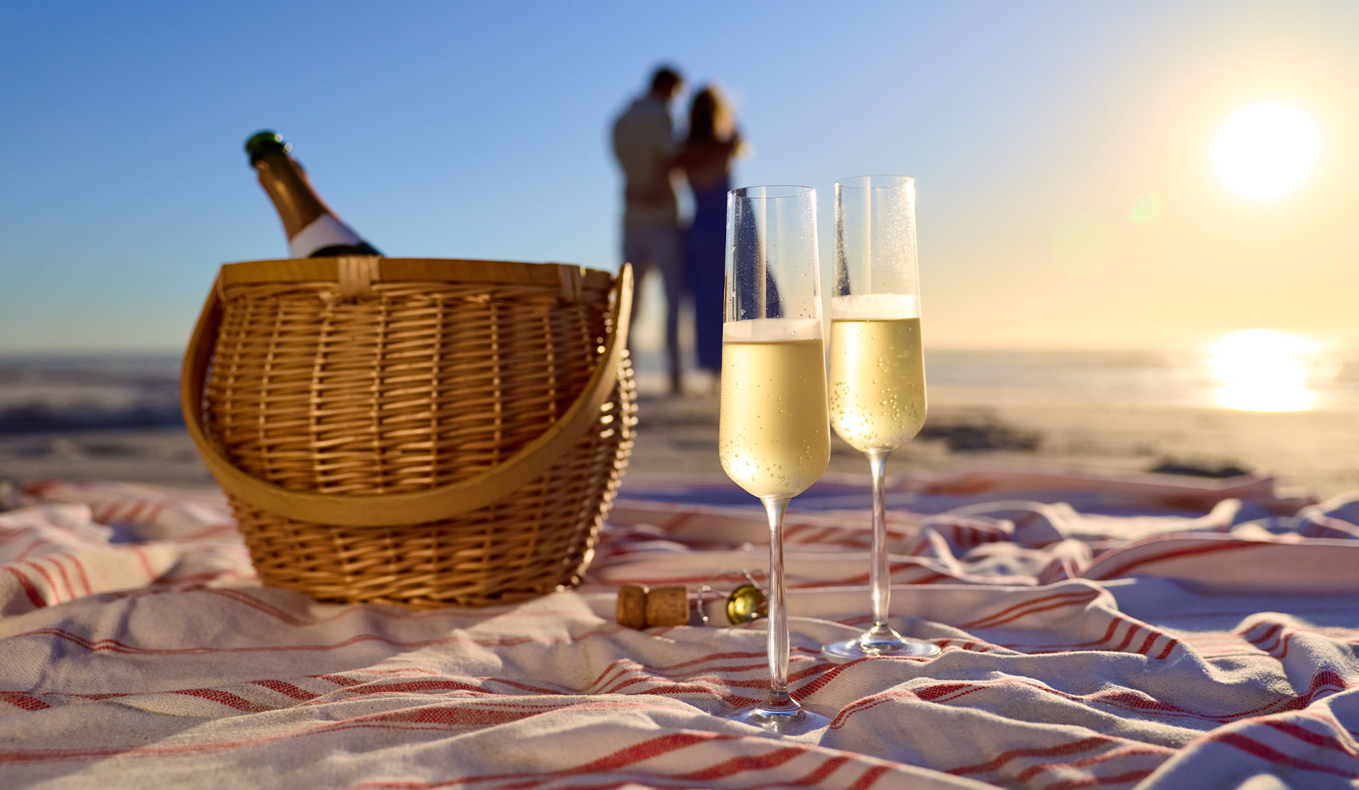 Picknick am Strand mit Sektflasche, Gläsern und einem Paar im Hintergrund bei Sonnenuntergang.