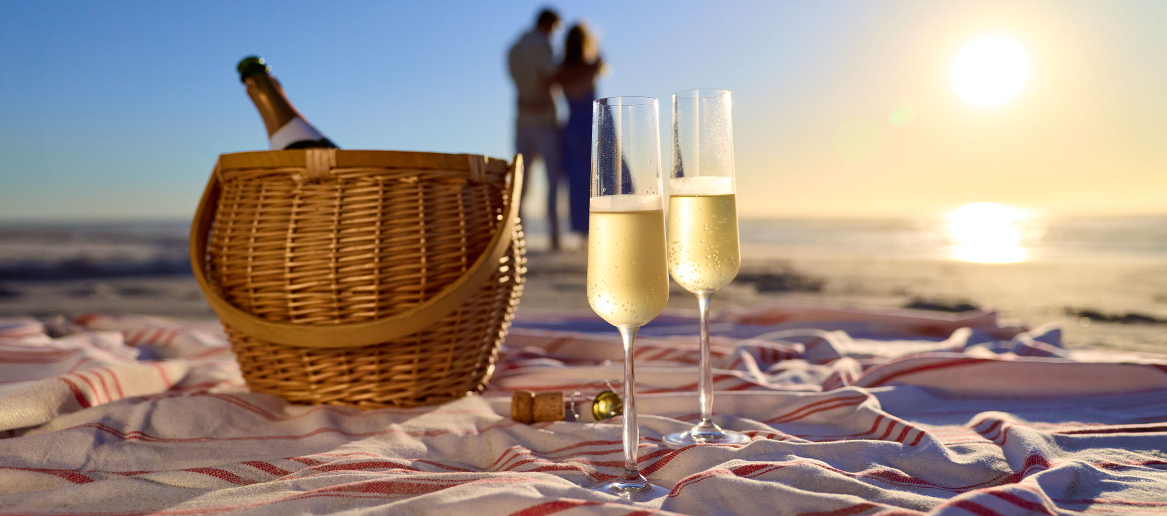 Sektgläser und Picknickkorb auf einer Stranddecke mit einem Paar im Hintergrund.