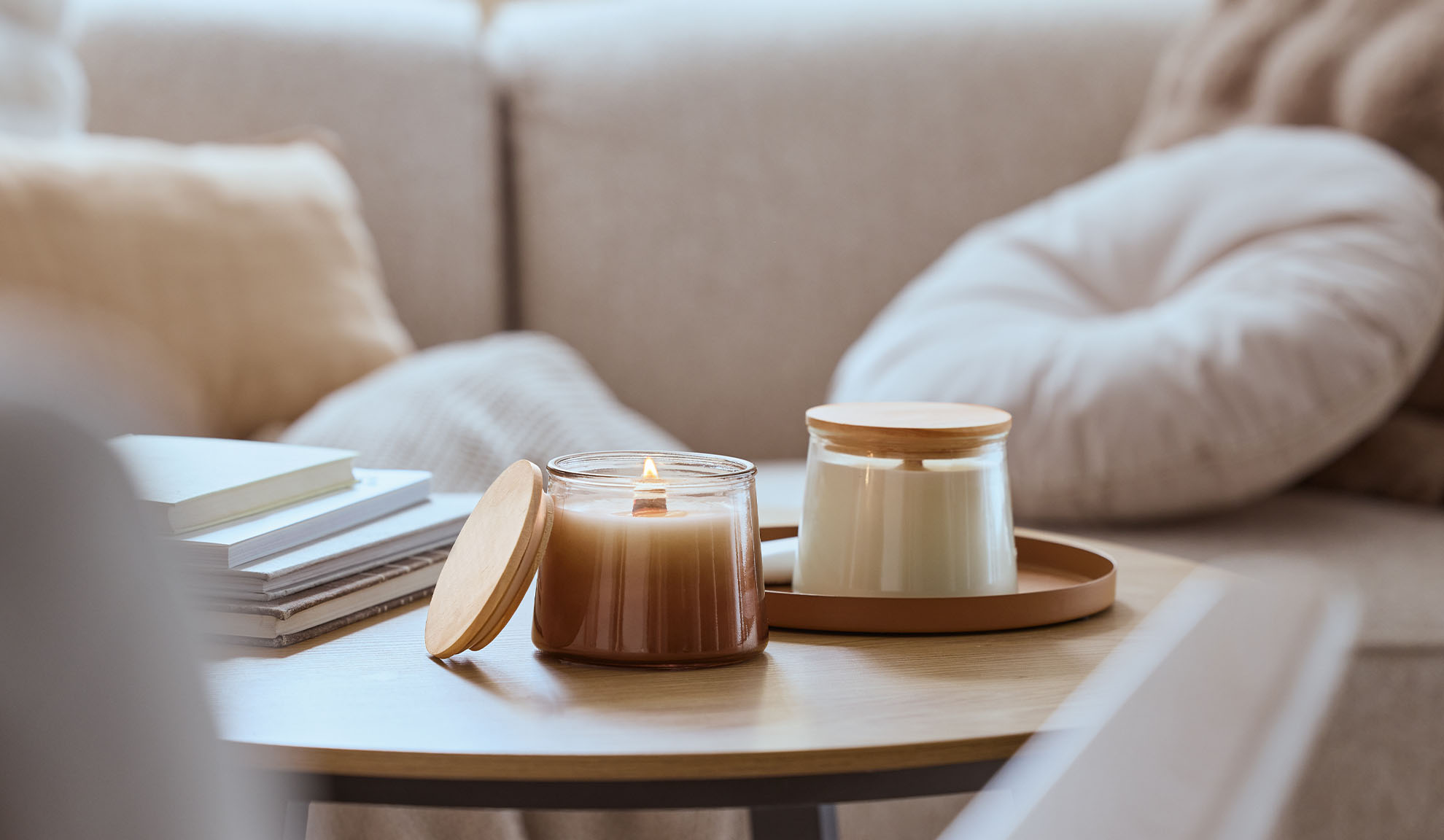 Zwei Duftkerzen auf einem Holztisch, eine brennend, mit einem gemütlichen Sofa im Hintergrund.