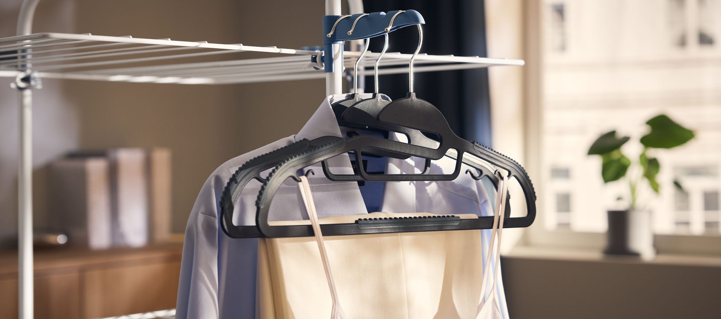 Kleidung auf Kleiderbügeln an einem Wäscheständer, mit einer Pflanze im Hintergrund.