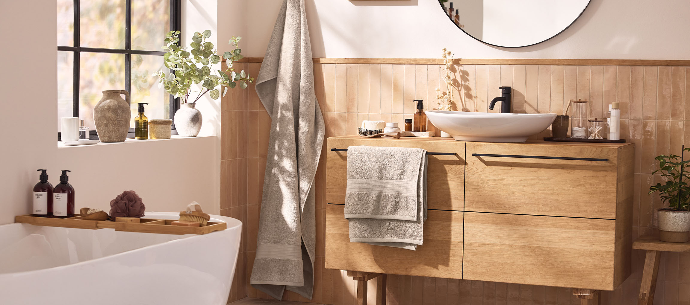 Ein modernes Badezimmer mit Holzwaschtisch, weißem Waschbecken, rundem Spiegel und Badewanne mit Badaccessoires.