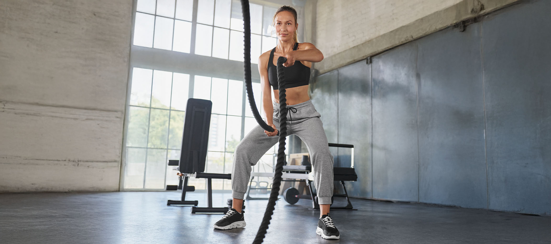 Frau in schwarzem Sport-BH und grauer Jogginghose trainiert mit Battle Ropes