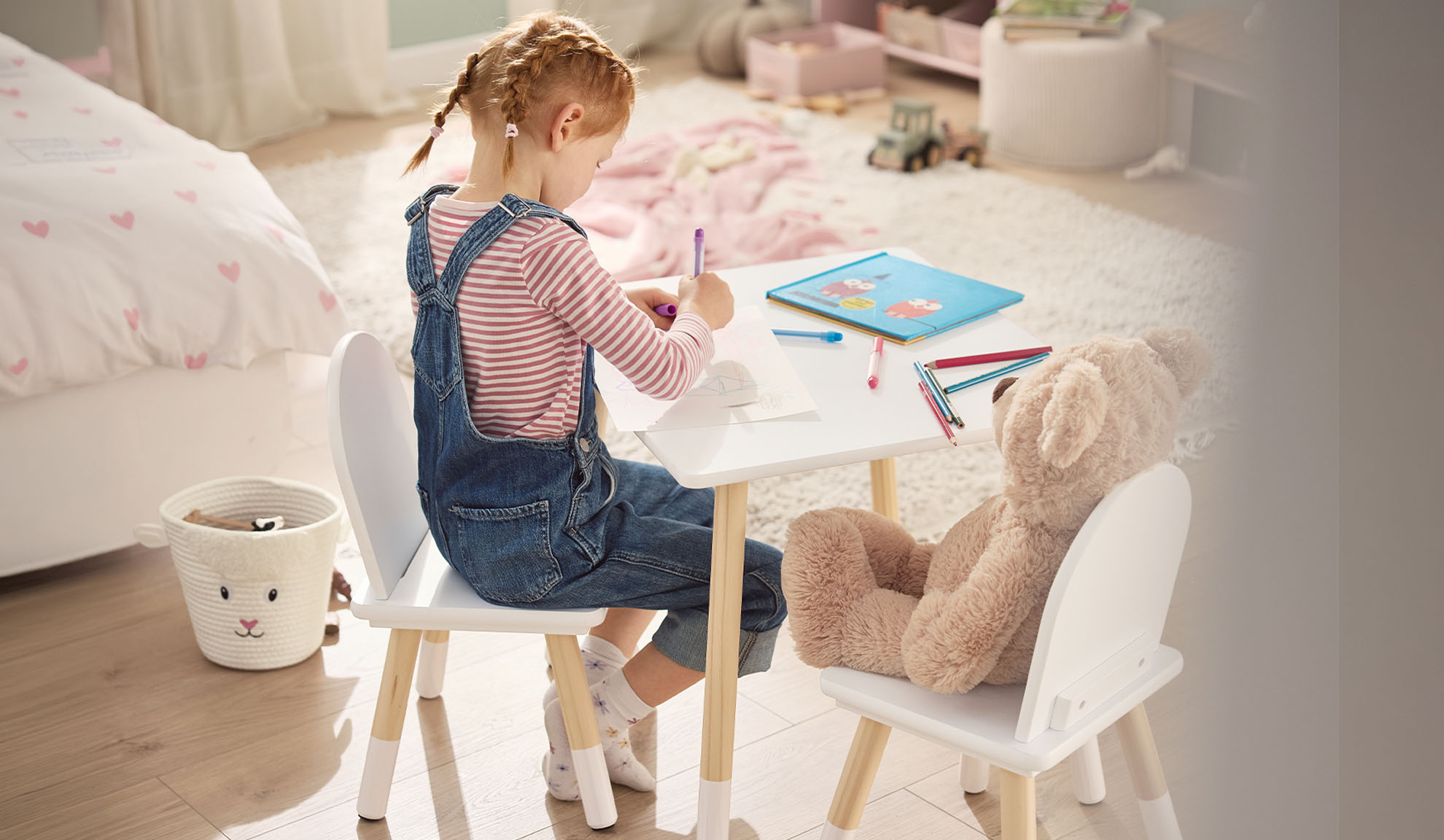 Mädchen zeichnet an einem weißen Tisch mit einem Teddybär, im Kinderzimmer.