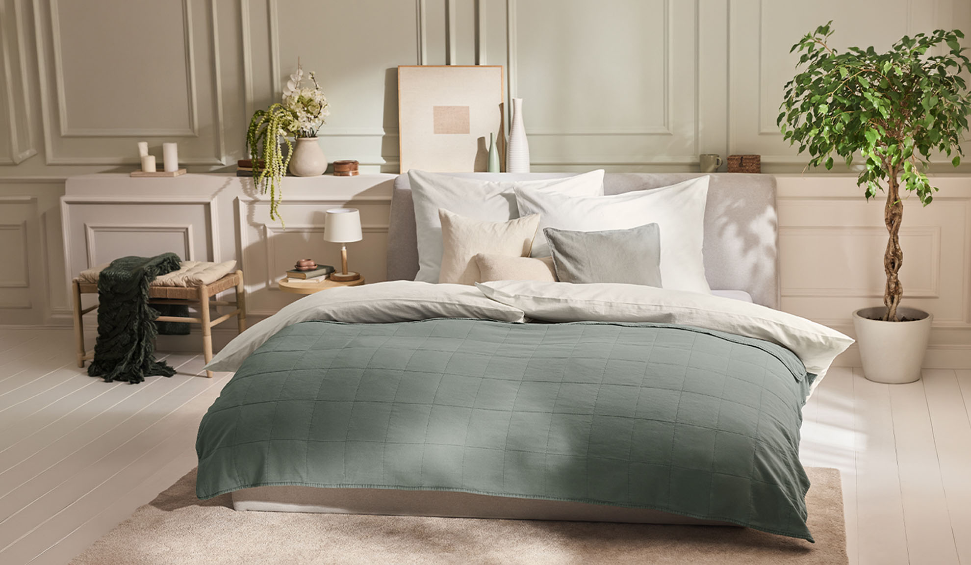 Schlafzimmer mit Bett, grünem Bettbezug, weißen Kissen und einer Pflanze
