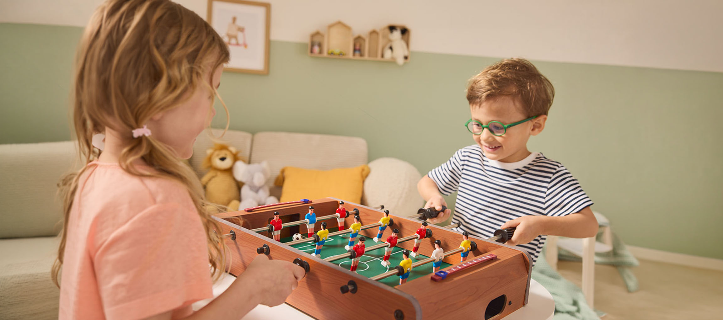 Zwei Kinder spielen Tischfußball, ein Mädchen in einem rosa Hemd und ein Junge in einem gestreiften Hemd.
