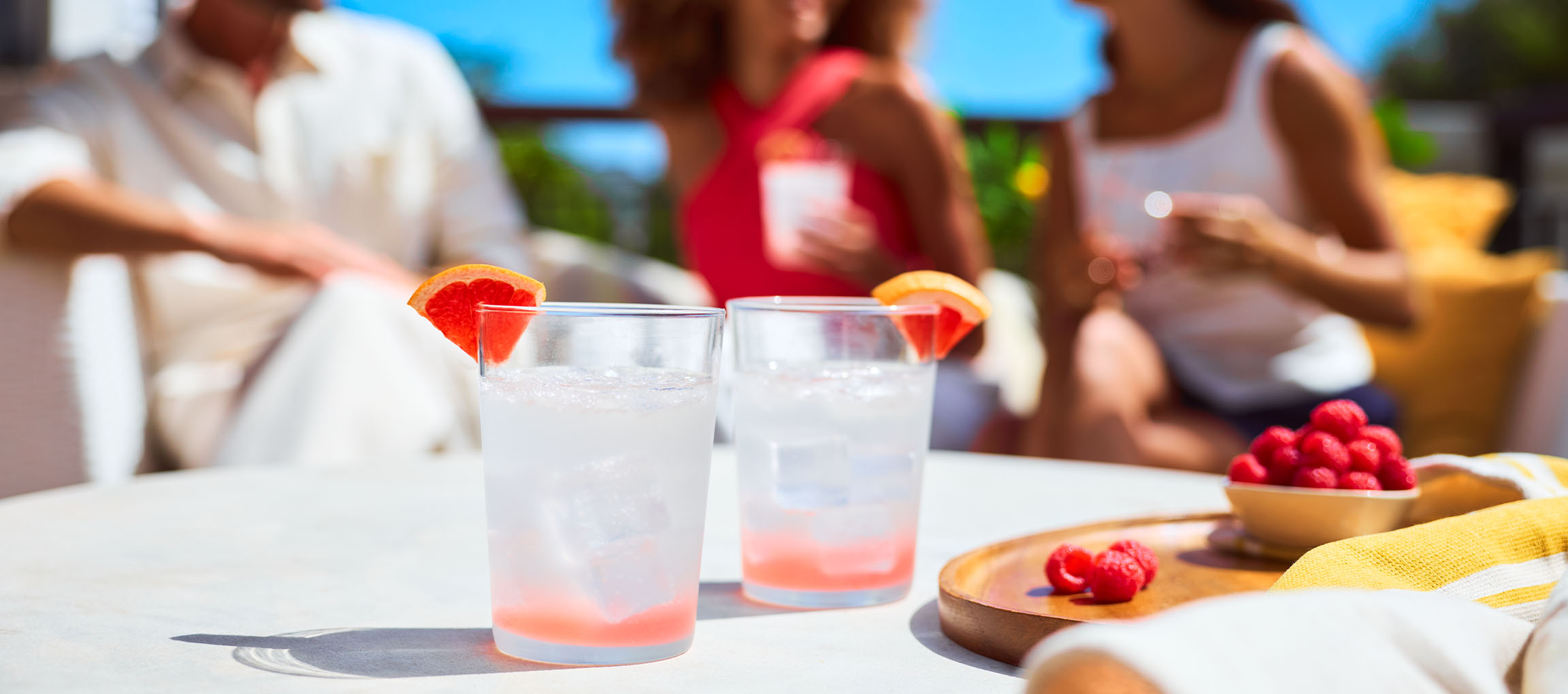 Erfrischende Grapefruitgetränke und Himbeeren auf einem sonnigen Tisch im Freien mit entspannten Menschen.