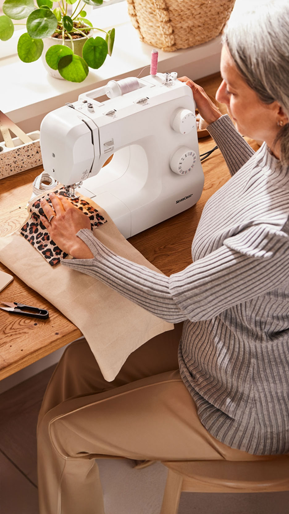 Señora mayor cosiendo unas telas de colores tierra con una máquina de coser blanca y con hilo de color sobre mesa de costura
