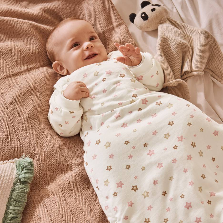 Lächelndes Baby in weißem Schlafsack mit rosa und braunen Sternen, auf einer Decke liegend.