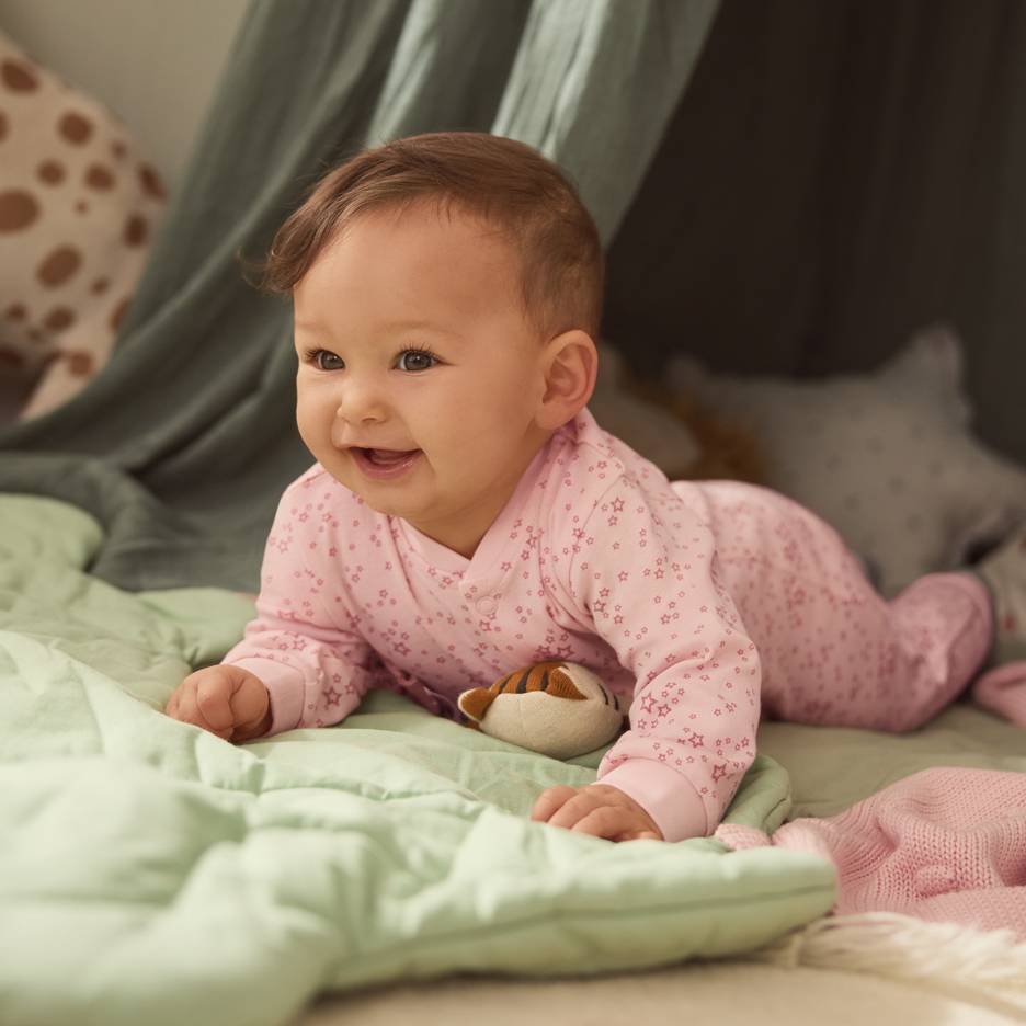 Baby in rosa Sternenpyjama, auf einer grünen Decke mit einem Plüschtier liegend.
