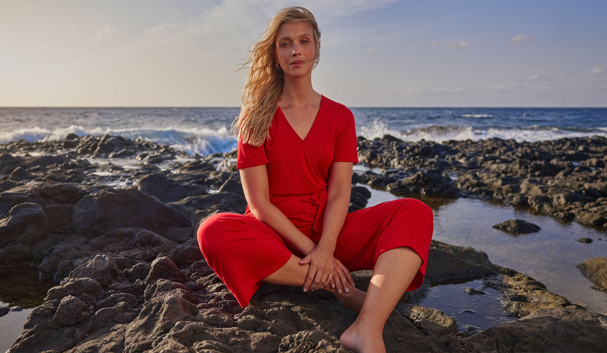 Frau in rotem Overall sitzt auf Felsen am Meer.