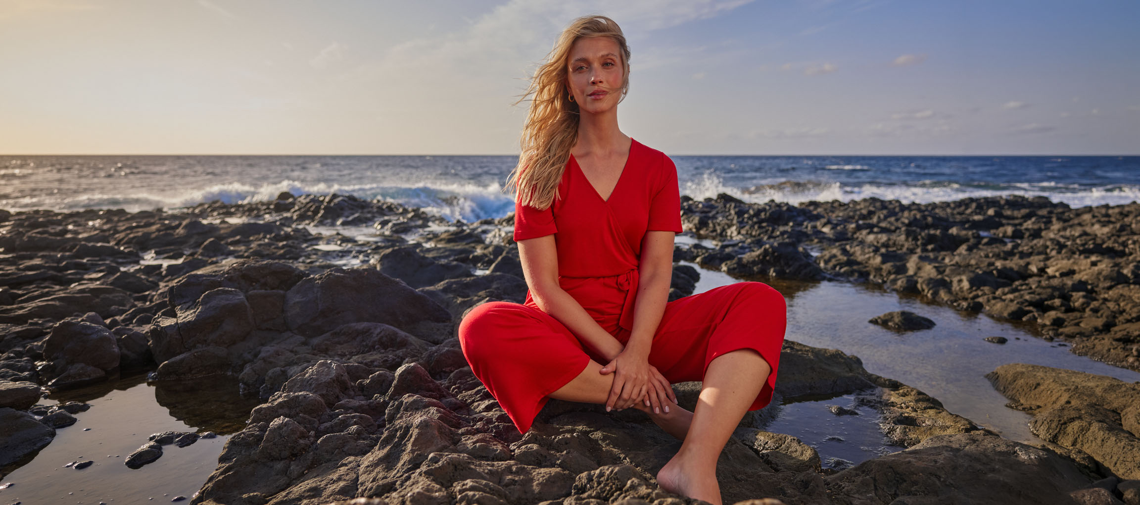 Frau in rotem Overall sitzt an einem felsigen Strand am Meer.