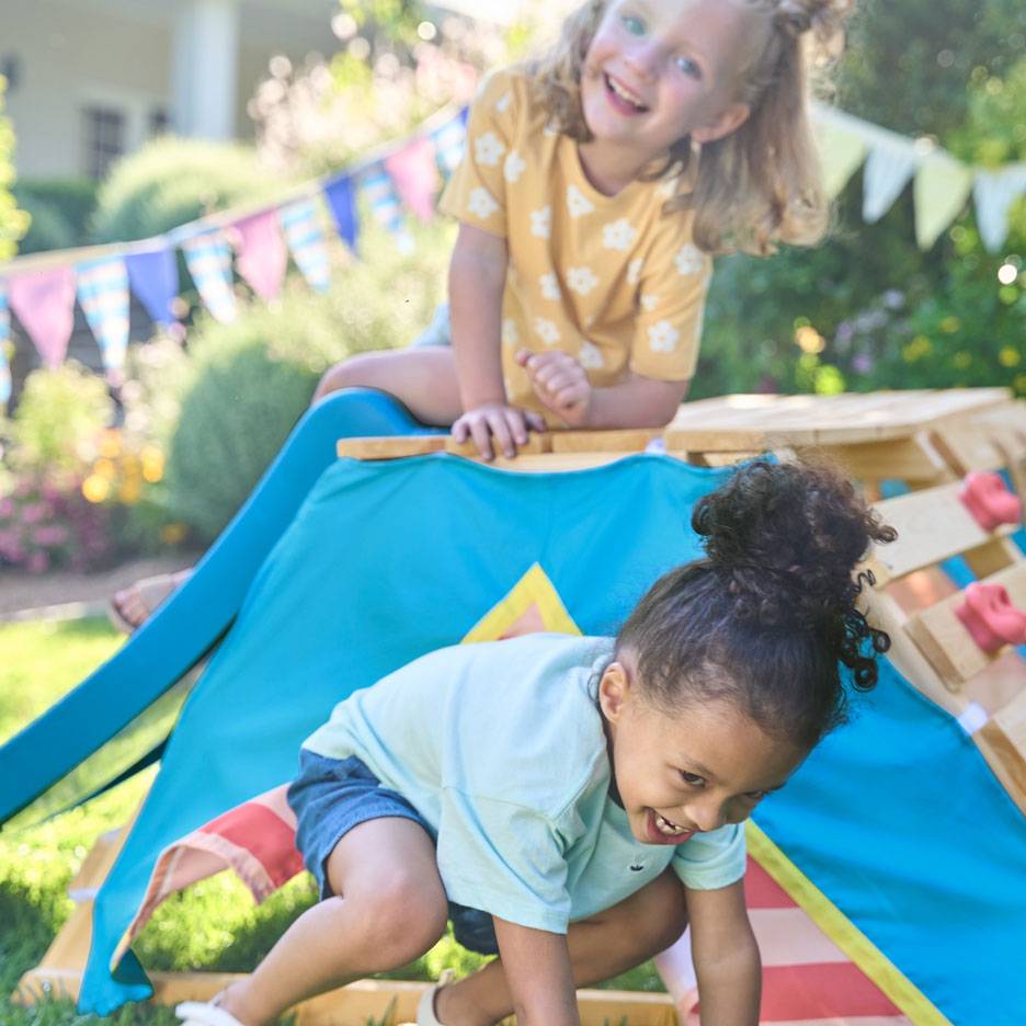 Zwei glückliche Kinder spielen auf einem hölzernen Klettergerüst mit blauem Zelt in einem sonnigen Garten.