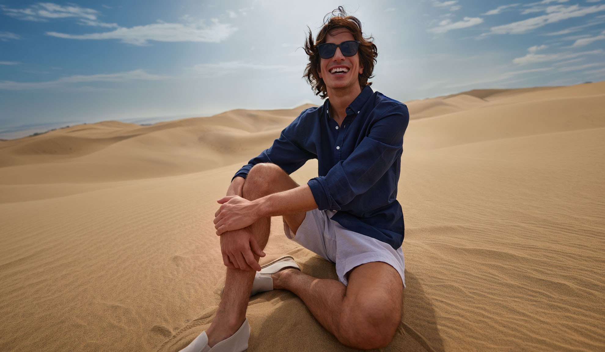 Mann in dunkelblauem Hemd und hellen Shorts sitzt lächelnd auf Sanddünen.