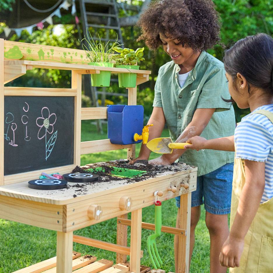 Zwei Kinder spielen mit einer Matschküche aus Holz, mit Kreidetafel und Topfpflanzen.