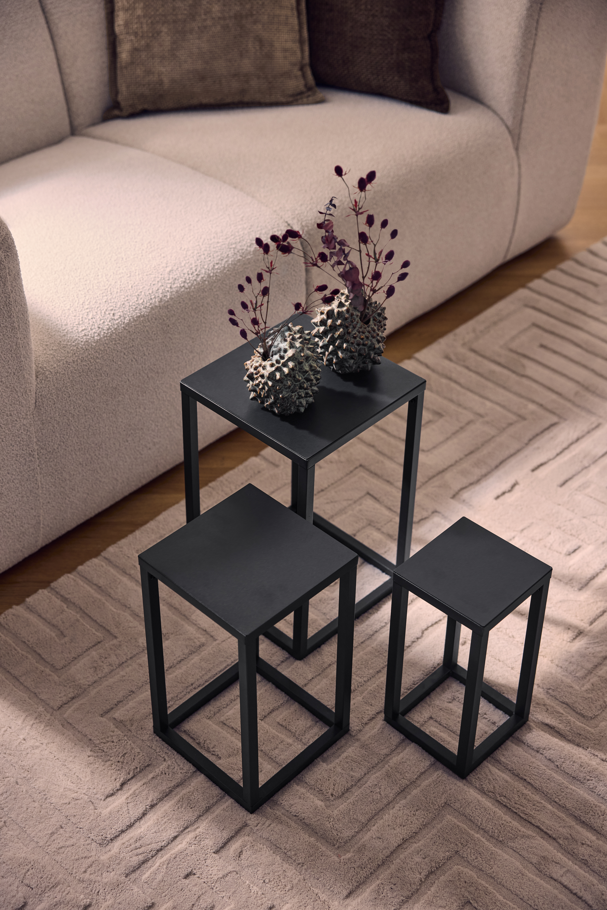 Drei schwarze Metall-Couchtische auf einem beigen Teppich mit geometrischem Muster, neben einem hellen Sofa.