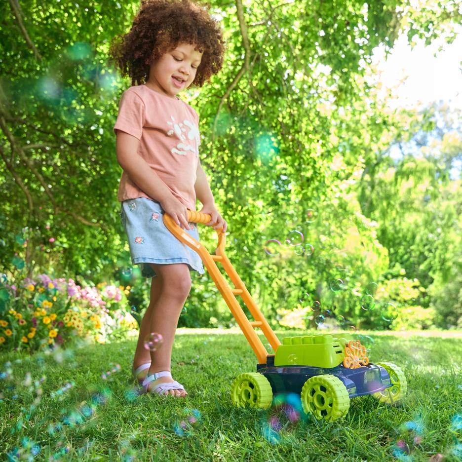 Ein Kind mit lockigem Haar schiebt einen Spielzeug-Blasenrasenmäher auf grünem Gras.