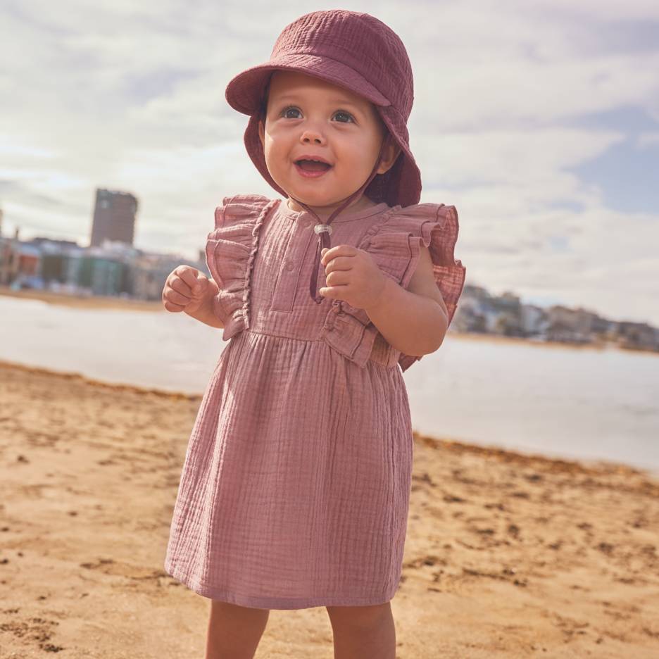 Baby in rosa Kleid und Hut am Strand.