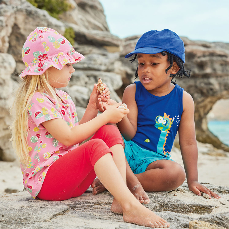 Zwei Kinder am Strand, eines mit rosa Sonnenhut und Obstmuster-Oberteil, das andere mit blauer Kappe und Dinosaurier-Tanktop, Muscheln haltend.
