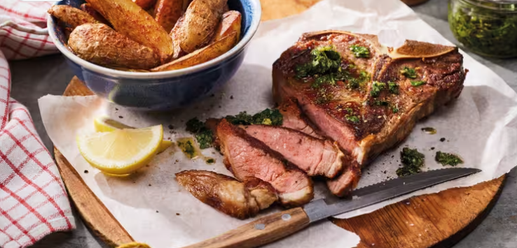 T-Bone-Steak mit Kräutersauce, Zitronenspalten und Kartoffelspalten auf einem Holzbrett.