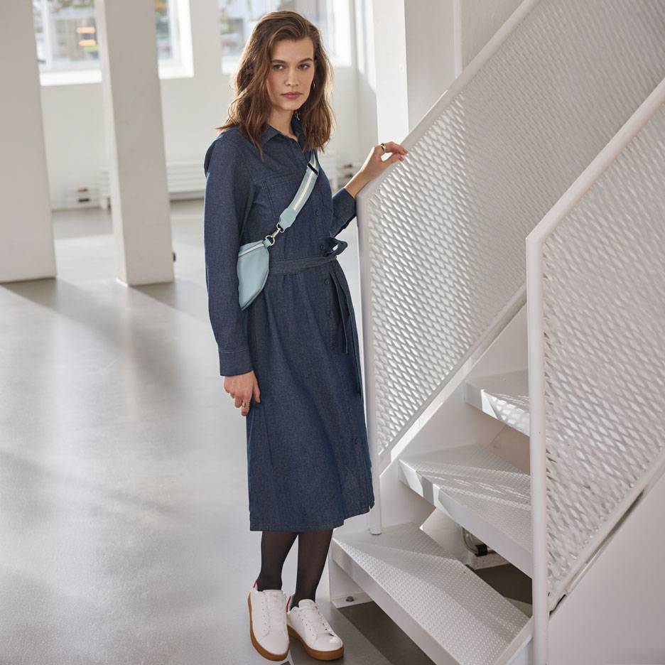 Frau in Jeanshemdkleid, schwarzer Strumpfhose und weißen Sneakern, mit hellblauer Bauchtasche.