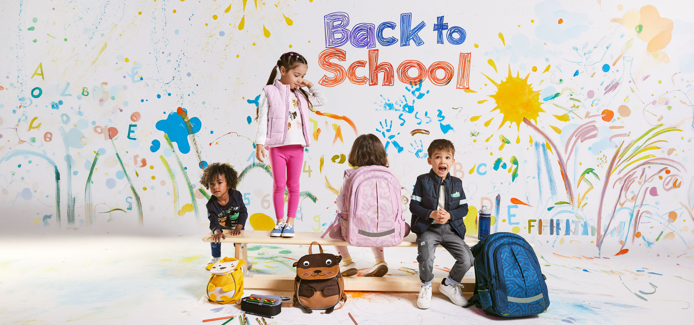 Kinder mit Rucksäcken und Schulmaterial vor einem Hintergrund bunter Zeichnungen mit dem Text