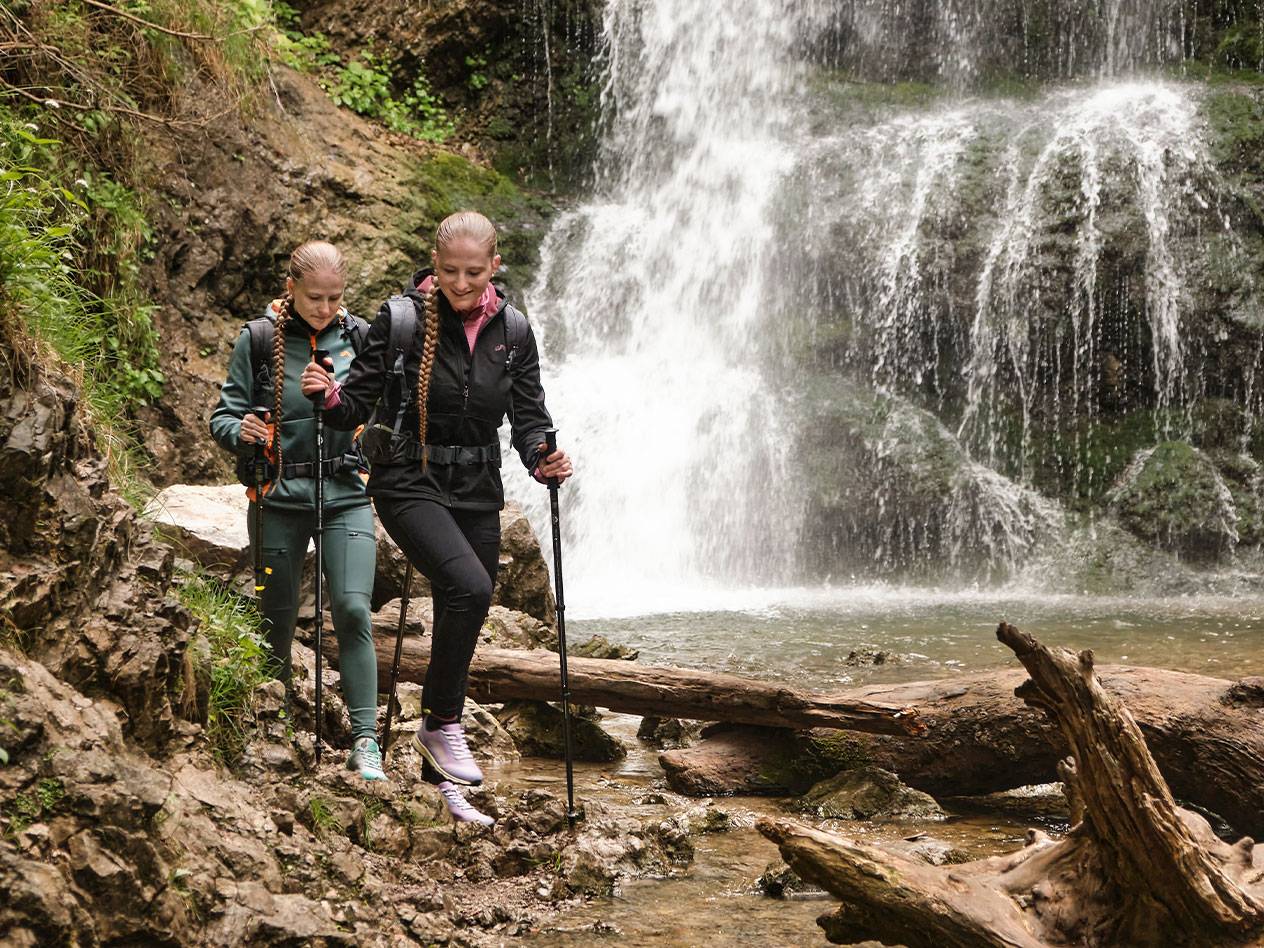 Zwei Frauen mit Trekkingstöcken wandern einen felsigen Pfad entlang in der Nähe eines Wasserfalls.