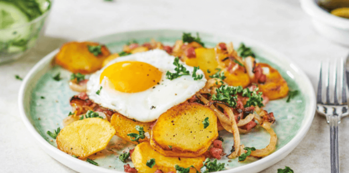 Spiegelei auf Bratkartoffeln mit Zwiebeln, Speck und Petersilie auf einem Teller.